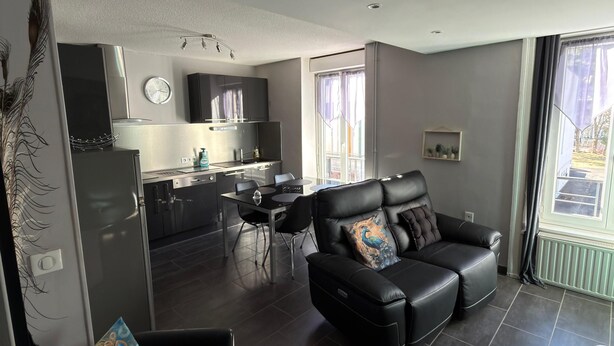 Ein modernes Wohnzimmer mit einem schwarzen Ledersofa und einem weißen Sessel, ein Essbereich mit Tisch und Stühlen sowie eine Küche im Hintergrund.