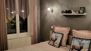 Ein Schlafzimmer mit einem Bett, zwei Zierkissen, einem Nachttisch, einer Wandablage und einem Fenster mit Vorhängen.