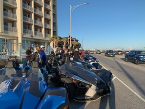 A row of parked motorcycles on a street with a multi-story building in the background.