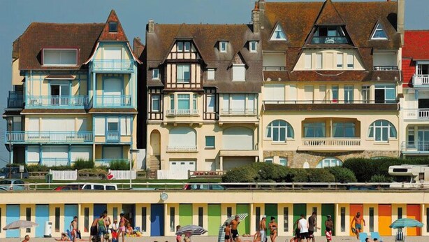 Une rangée de bâtiments colorés situés en bord de mer, dotés de balcons, une plage au premier plan et des personnes qui prennent un bain de soleil.