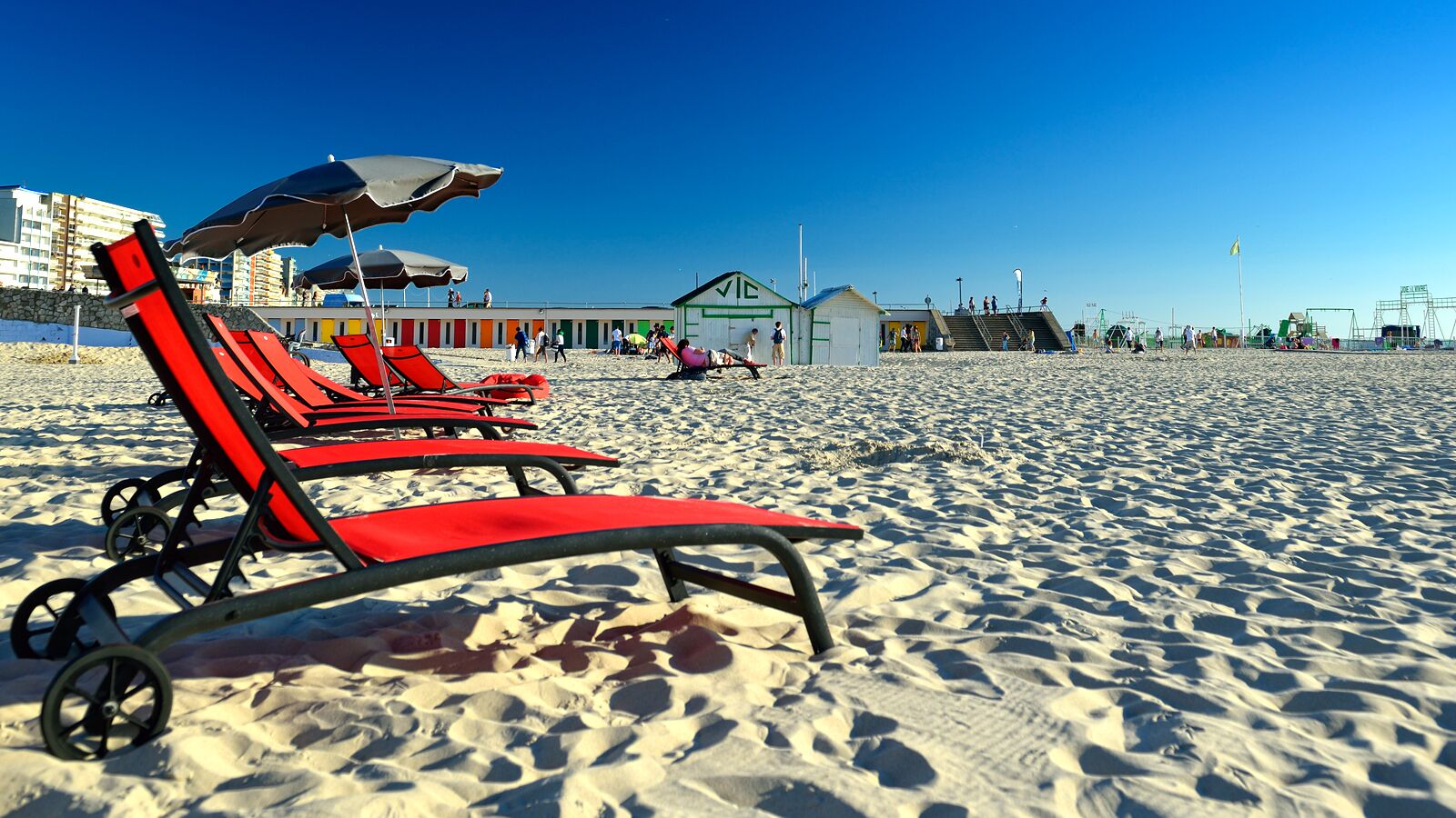 Une plage avec des chaises longues et des parasols rouges, une tour de surveillance et, au fond, un bâtiment doté d’un toit végétalisé.
