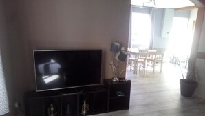 Un salon avec un téléviseur à écran plat posé sur un meuble noir, un coin repas avec une table et des chaises en bois, et une plante en pot.