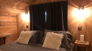 Quarto de cabana de madeira com uma cama, duas almofadas macias, um criado-mudo com uma vela e uma janela com cortinas.