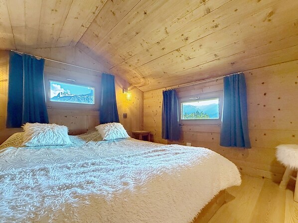 A wooden cabin bedroom with a bed, two windows with curtains, and a view of the mountains. - Small chalet with breathtaking views of Mont-Blanc, 5 minutes from the ski slopes (Sallanches)