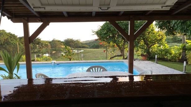 A covered outdoor area with a pool, chairs, and a view of a garden and hills.