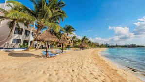 A beach with palm trees, lounge chairs, and a building with a thatched roof. - Ground-Floor Beachfront Living with Private Patio (Akumal)