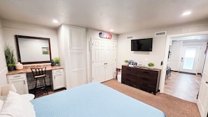A hotel room with a bed, a desk with a chair, a dresser, a TV mounted on the wall, and a door leading to another room.