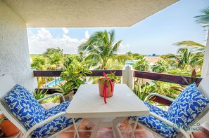 Un balcon avec une table blanche et des chaises à motifs bleus, donnant sur un jardin luxuriant, avec des palmiers et un ciel dégagé.