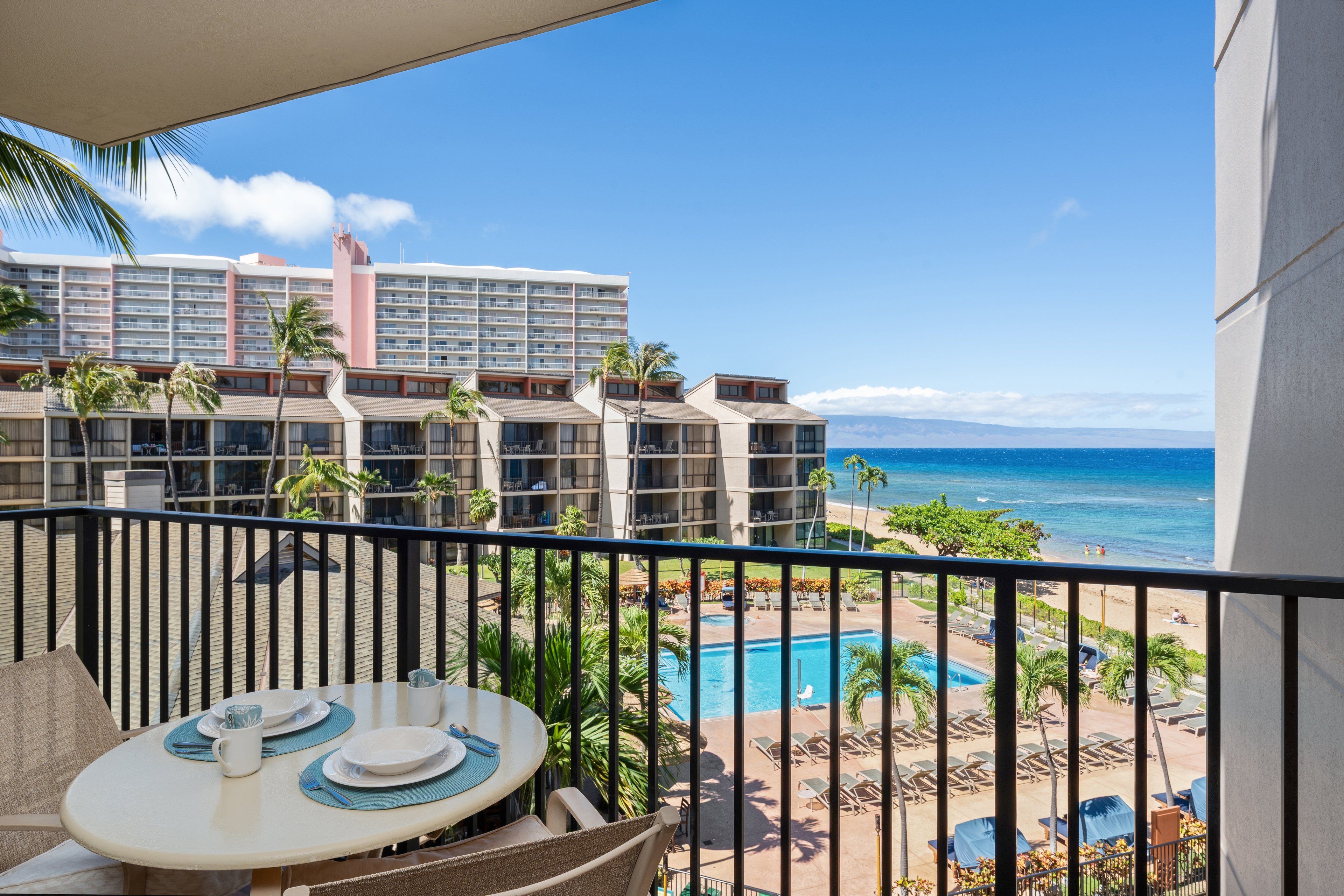 A balcony with a table set for dining, overlooking a swimming pool and beachfront.