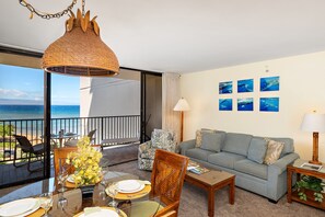 A room with a balcony overlooking the sea, a dining area with a table set for two, a sofa, and a wall with framed pictures.