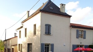 Een gebouw met twee verdiepingen, een witte gevel, meerdere ramen met luiken en een grijze hek.