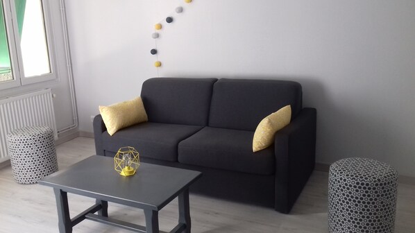 A modern living room with a dark grey sofa, two patterned ottomans, a small table with a geometric glass sculpture, and a string of decorative balls on the wall.