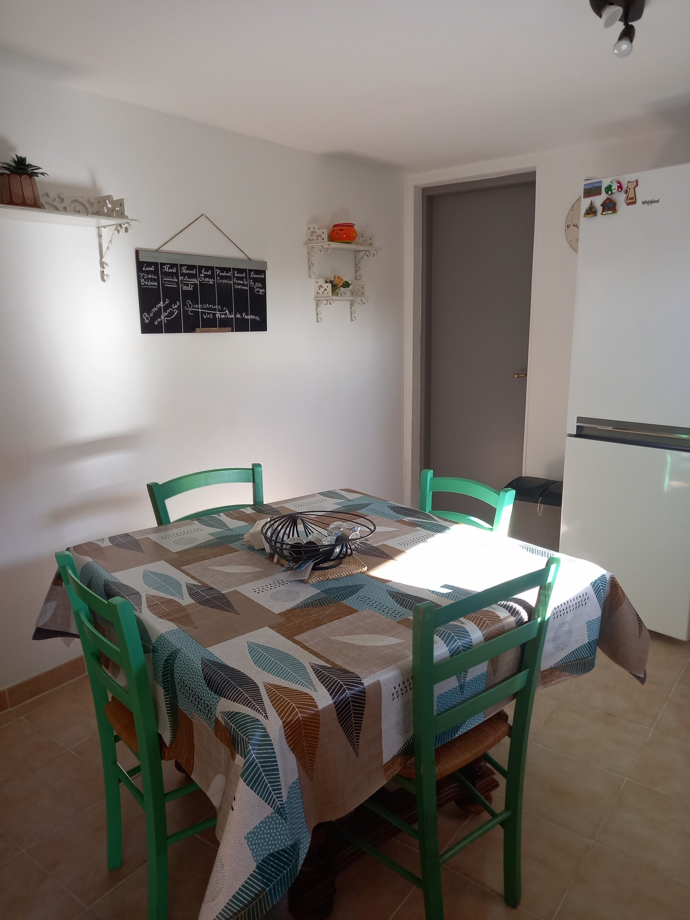 A dining area with a table covered by a patterned tablecloth, surrounded by green chairs. A blackboard with handwritten notes is mounted on the wall above the table.