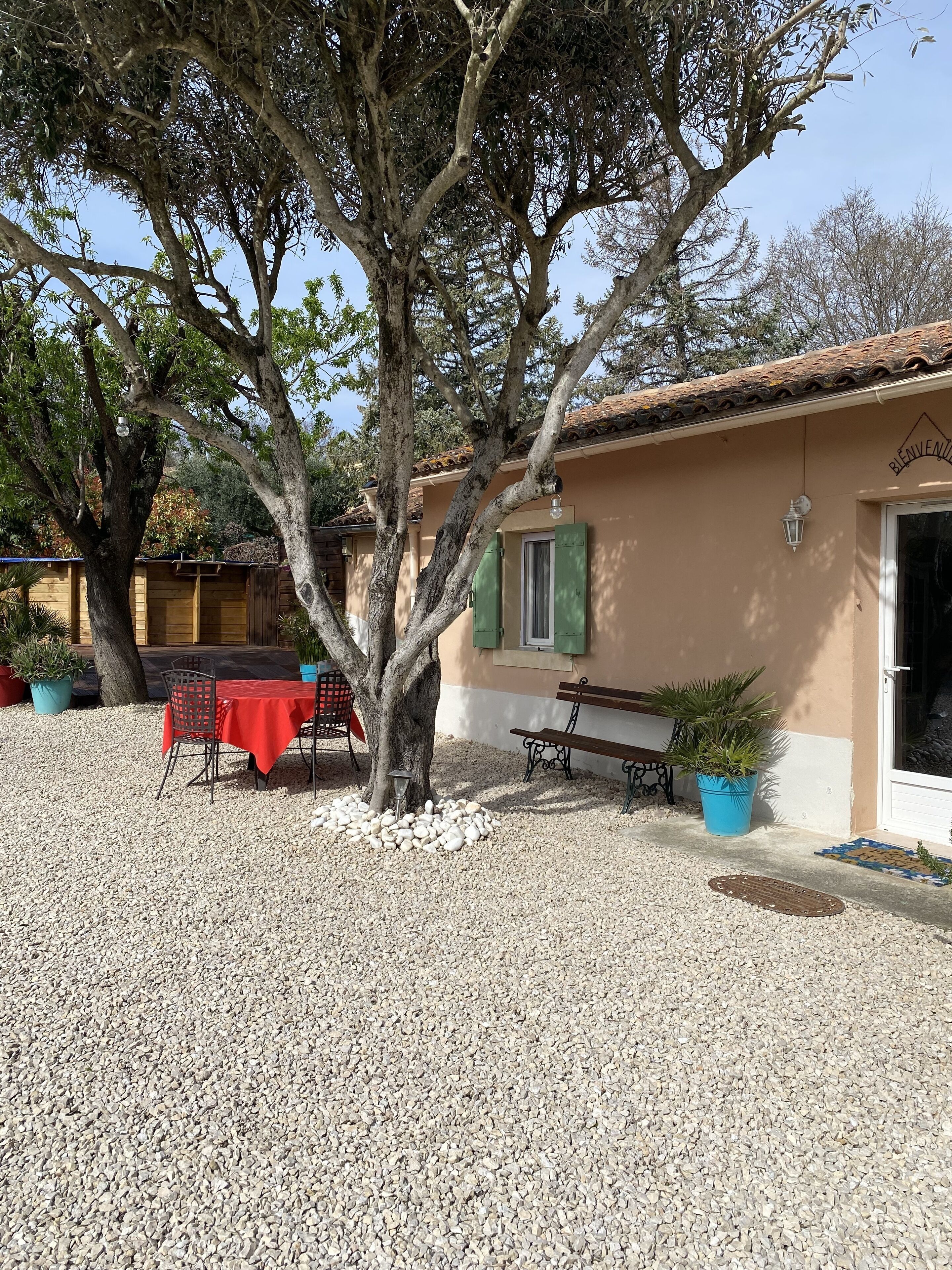 A building with a beige exterior, a red table with chairs, a bench, and potted plants.