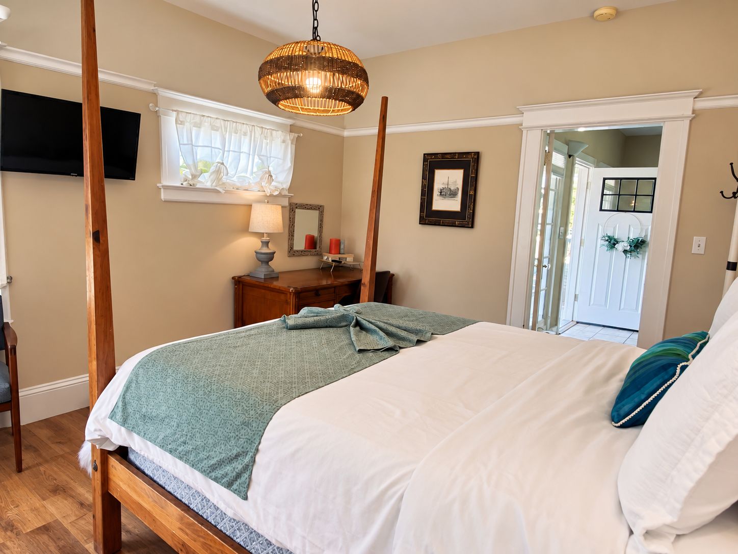 A bedroom with a four-poster bed, a bedside table with a lamp, a framed picture on the wall, and a door leading to another room.