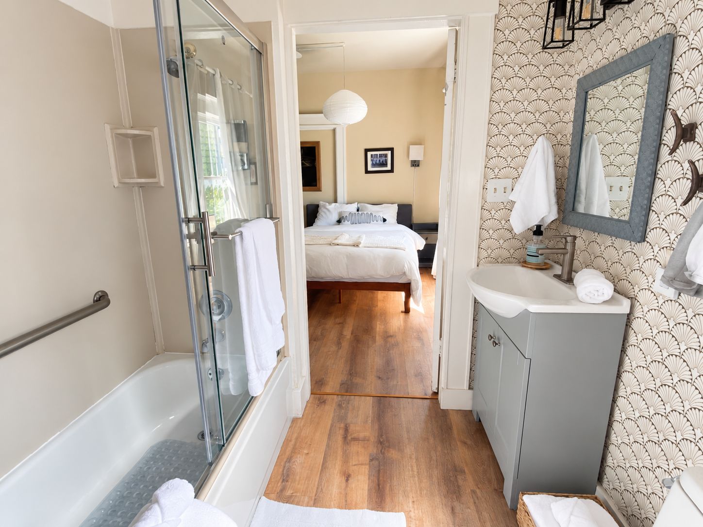 Une salle de bain d’hôtel avec une douche à parois de verre, un plancher en bois, un lavabo avec un miroir et un lit visible à travers une porte ouverte.