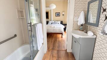 Une salle de bain d’hôtel avec une douche à parois de verre, un plancher en bois, un lavabo avec un miroir et un lit visible à travers une porte ouverte.