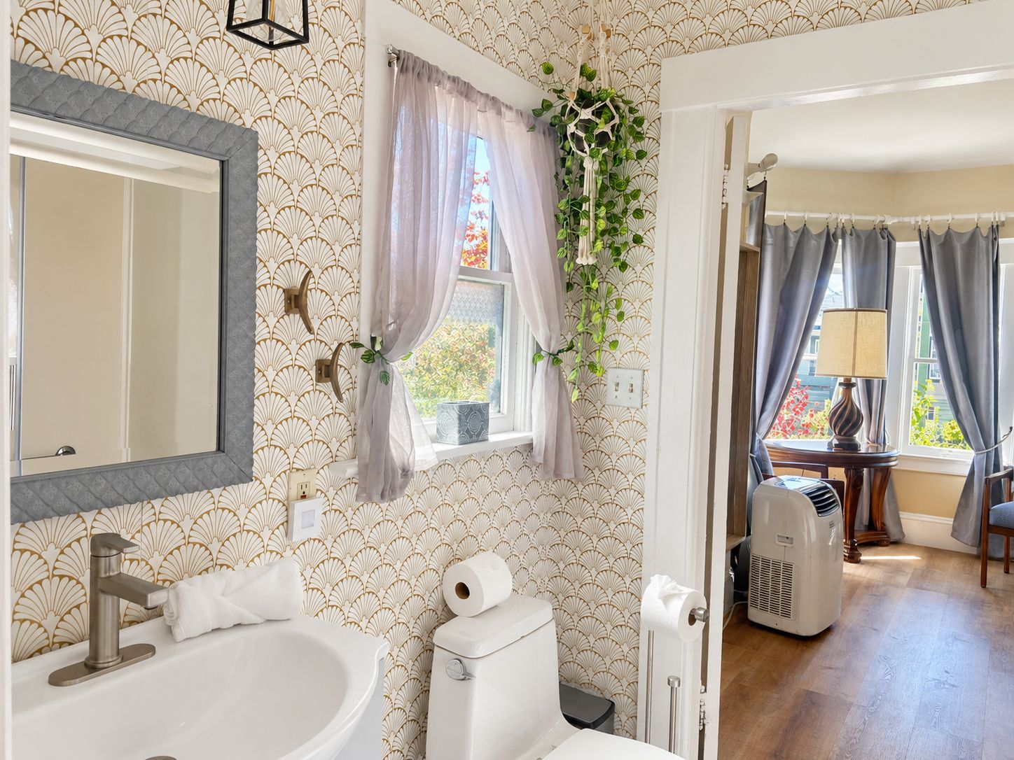 A bathroom with a sink, toilet, and mirror. There is a window with curtains and a plant hanging on the wall.