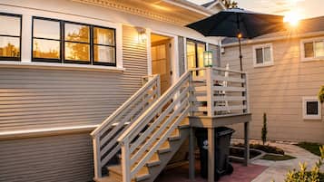 A residential building with a wooden staircase leading to a covered outdoor area, a trash bin, and a small garden.
