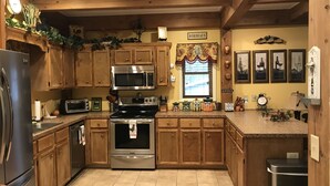 A kitchen with wooden cabinets, stainless steel appliances, and a window with a floral curtain. - Cedar Suite: Mountain Getaway overlooking The Roaring River on the vineyard (Traphill)