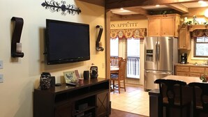 A spacious living room with a large flat-screen TV, wooden entertainment unit, and a glass coffee table. A kitchen area with a refrigerator and dining table is visible in the background. - Cedar Suite: Mountain Getaway overlooking The Roaring River on the vineyard (Traphill)