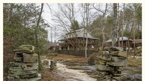 A rustic cabin with a stone foundation, surrounded by a forest. - Cedar Suite: Mountain Getaway overlooking The Roaring River on the vineyard (Traphill)