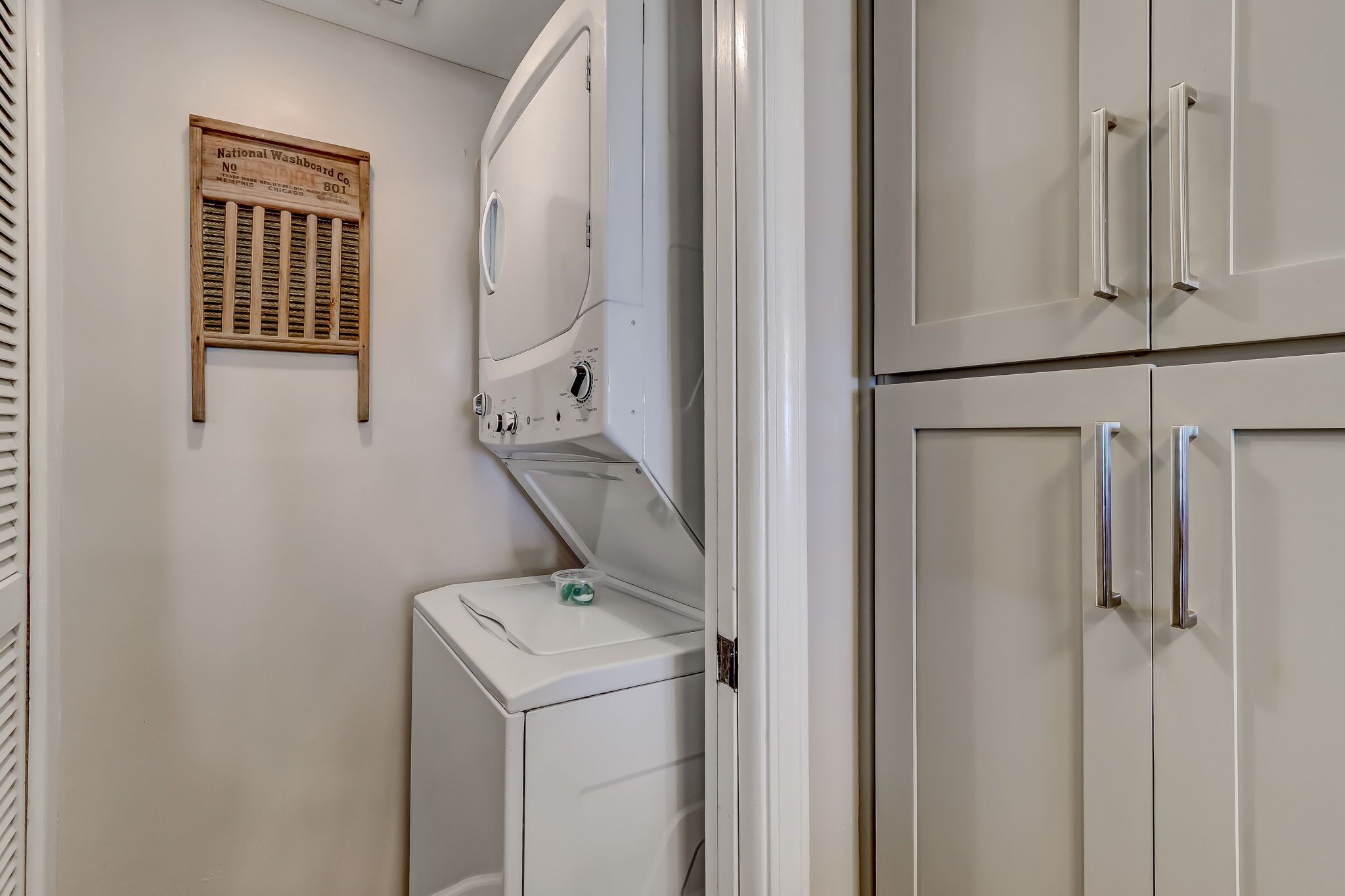 Une machine à laver blanche, avec une petite pancarte en bois fixée au-dessus, un petit récipient posé sur la machine, et une armoire blanche dotée de poignées argentées.