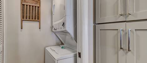Une machine à laver blanche, avec une petite pancarte en bois fixée au-dessus, un petit récipient posé sur la machine, et une armoire blanche dotée de poignées argentées.