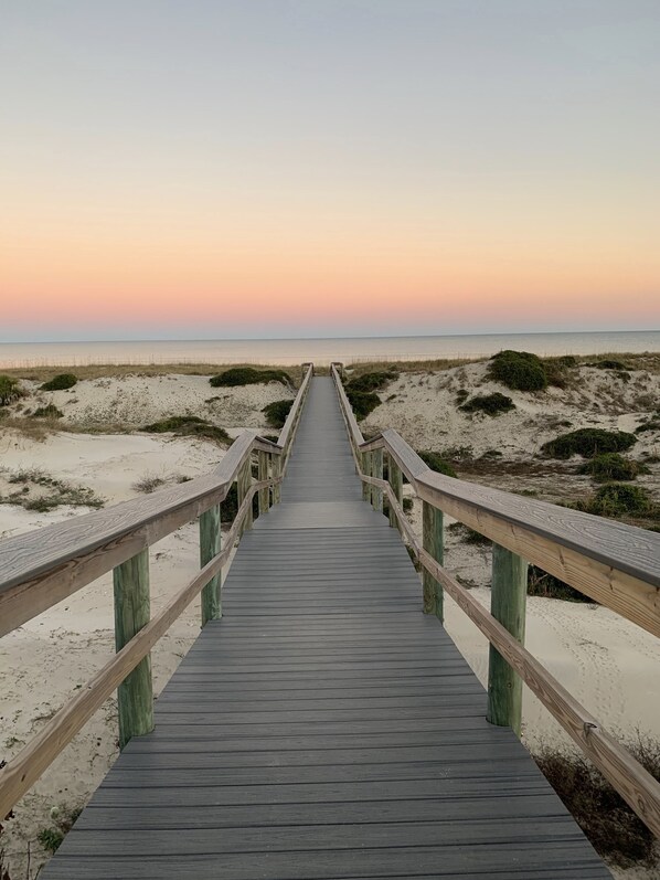 Un paseo marítimo de madera que lleva a una playa con dunas de arena y cielo despejado.