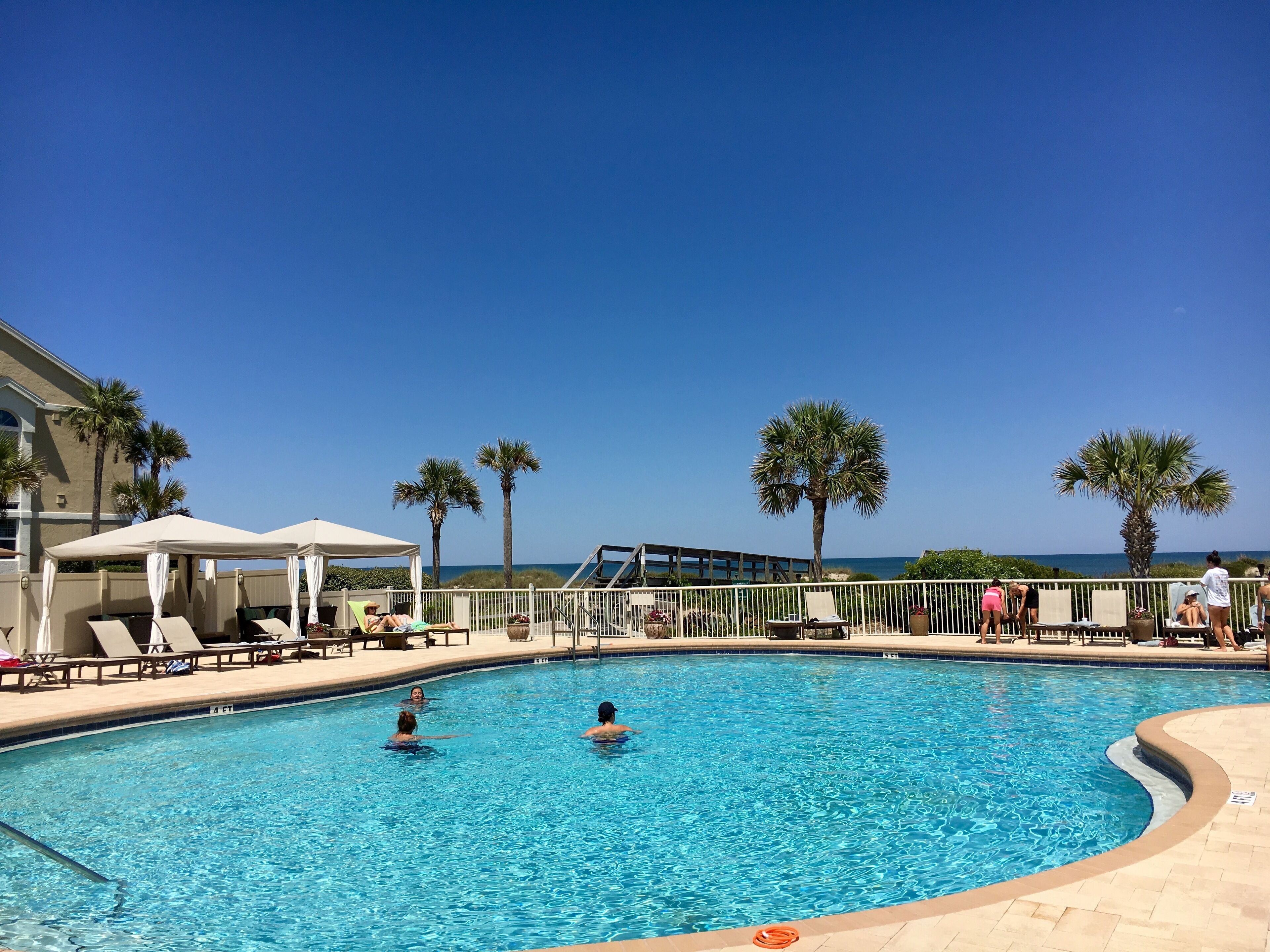 A swimming pool with lounge chairs and umbrellas, surrounded by palm trees and a clear sky.