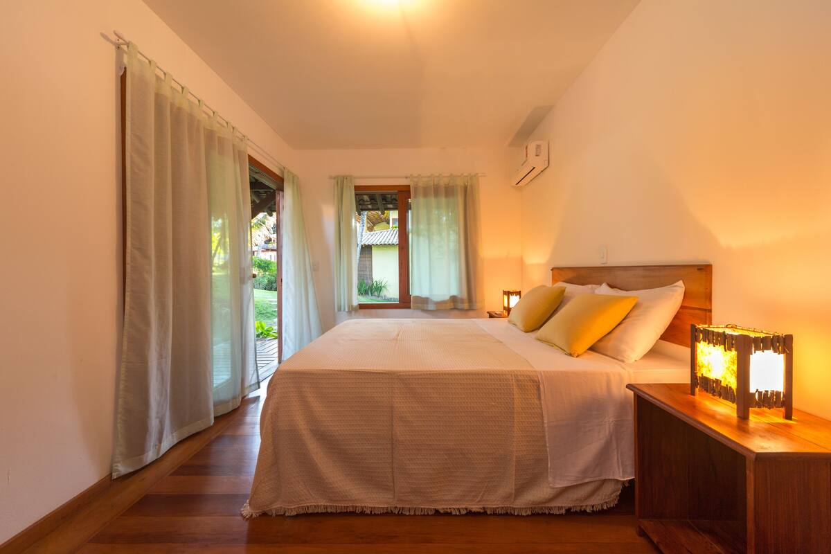 A bedroom with a large bed, a nightstand with a lamp, and a view of the outside through a window.