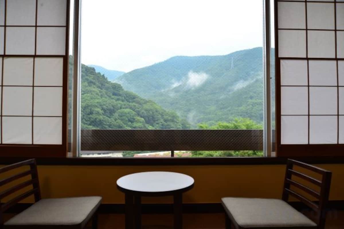 A room with a view of a mountainous landscape through a window with a grid pattern. There are two chairs and a small round table in the room.