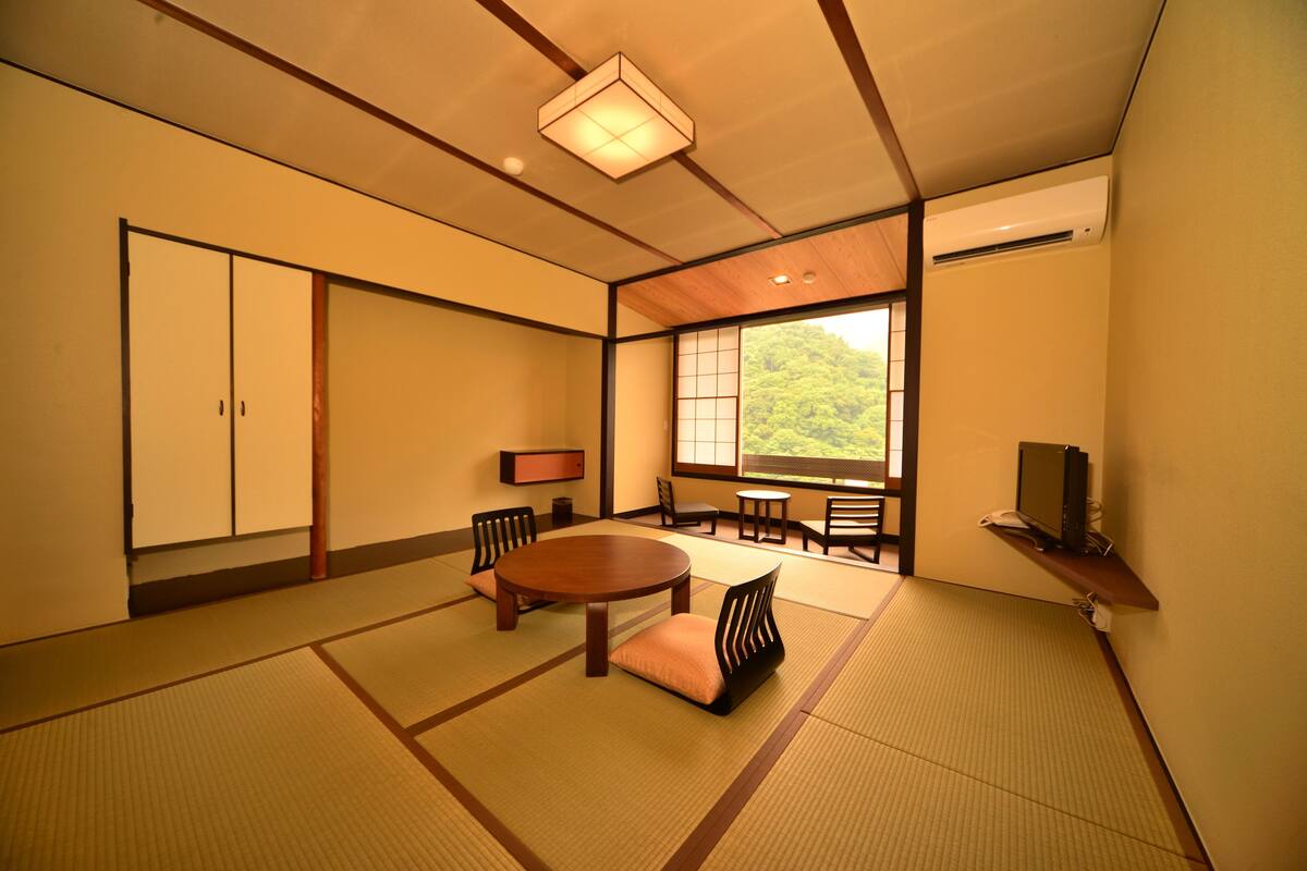 A traditional Japanese room with tatami flooring, a low table, chairs, a sliding door, and a window with a view of greenery.