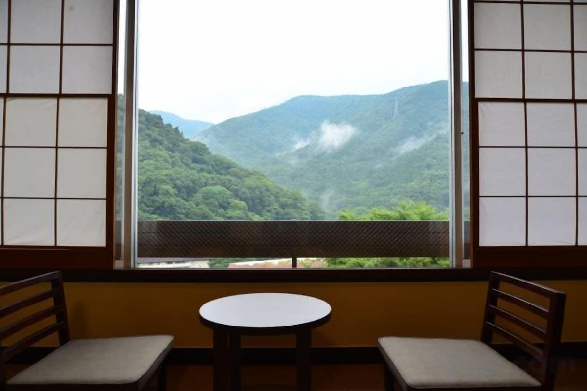 A room with a view of a mountainous landscape through a window with a grid pattern. There are two chairs and a small round table in the room.