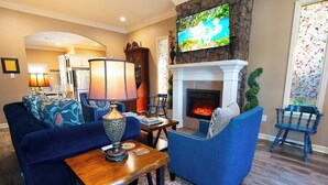 Una sala de estar con chimenea, sofás azules, una mesa con lámpara y un televisor montado en la pared.