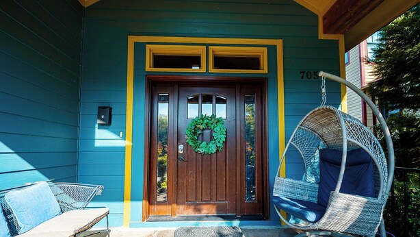 Puerta principal con un adorno de guirnalda, una silla colgante y un banco frente a un edificio con revestimiento azul y detalles amarillos.