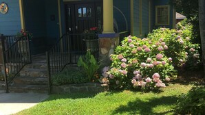 Una casa con porche delantero, columna de piedra y un jardín bien cuidado con flores rosadas.