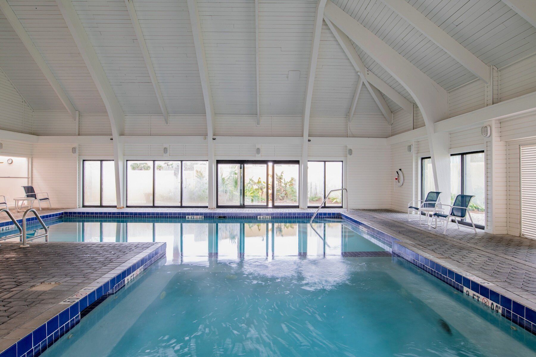 An indoor swimming pool with clear blue water, white walls, and large windows.