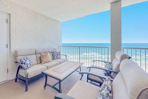 A balcony with a sofa and chairs, a table, and a view of the beach.