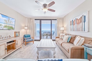 Un salon avec vue sur l’océan, un ventilateur de plafond, un téléviseur et une œuvre d’art sur le thème de la plage.