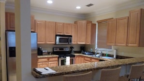 A kitchen with wooden cabinets, a microwave, stove, and a granite countertop. There are four wicker chairs at the bar.