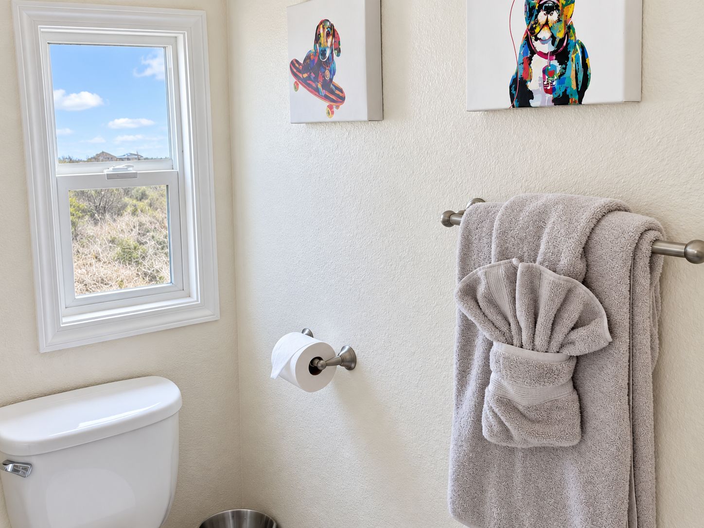 Een badkamer met een toilet, een raam met uitzicht op de omgeving en twee kunstwerken in lijsten aan de muur.