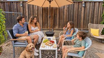 A family of four seated at an outdoor table with a variety of snacks and drinks, under a large umbrella, with a wooden fence in the background.