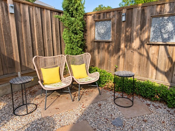 A patio area with two wicker chairs and two small round tables, surrounded by a wooden fence and decorative metal panels.