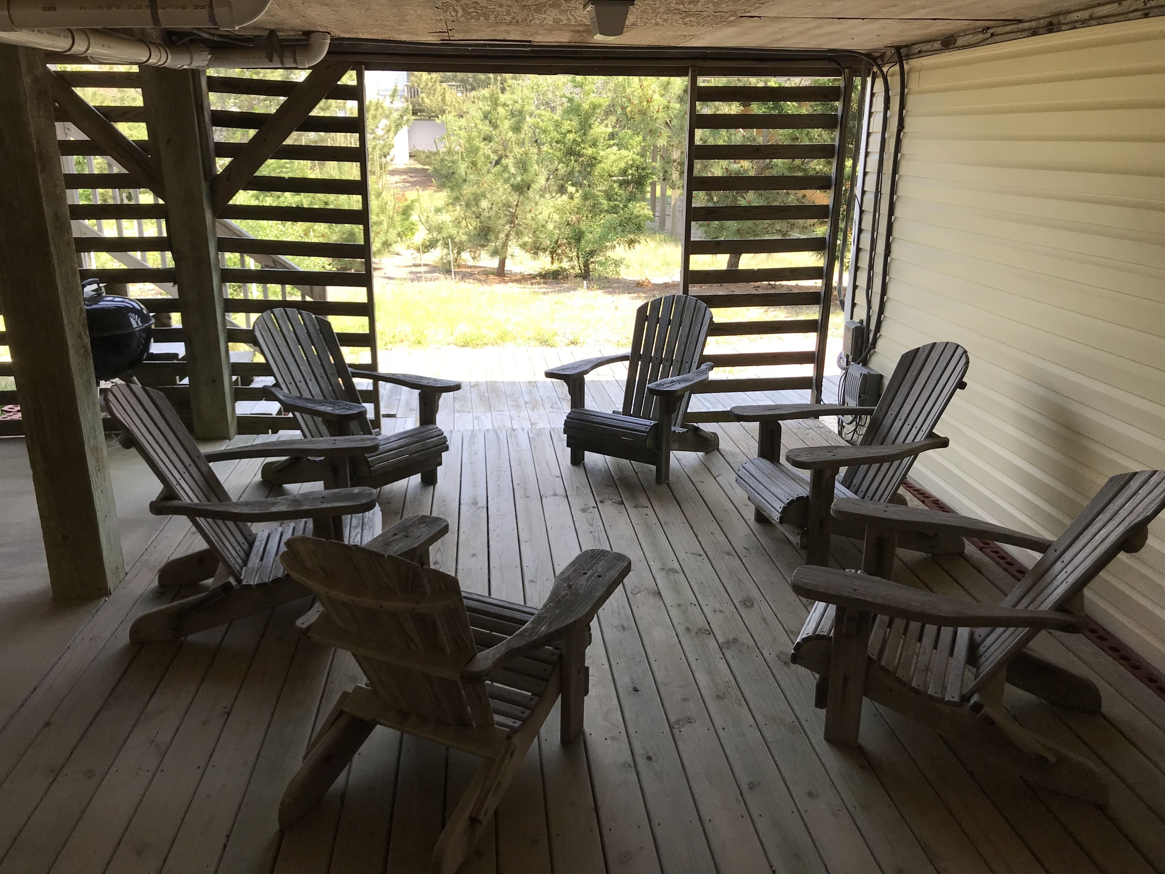 A covered outdoor seating area with wooden Adirondack chairs and a table, overlooking a garden with trees and a building in the background.