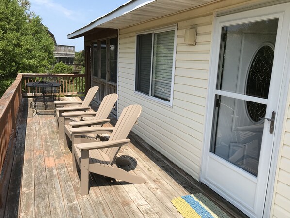 A wooden deck with Adirondack chairs and a table, a sliding glass door, and a view of a building in the background. - Clean & Cozy Beach Cottage Close to the Beach (Duck)