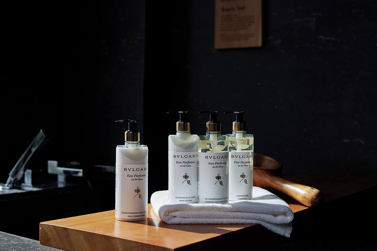 Un comptoir en bois sur lequel sont posées quatre bouteilles de parfum BVLGARI Eau de Parfum, une serviette blanche et une chaise en bois.