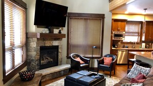A living room with a stone fireplace, a television mounted above it, and a kitchen area with wooden cabinets and a microwave. - Luxury Condo, Private Hot Tub, Perfectly Located (Frisco)