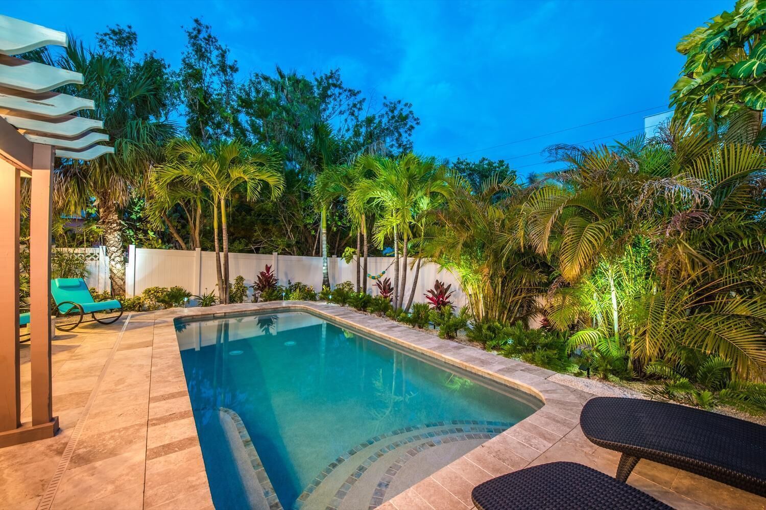 A swimming pool with a tiled deck, surrounded by lush tropical plants and a white wall.