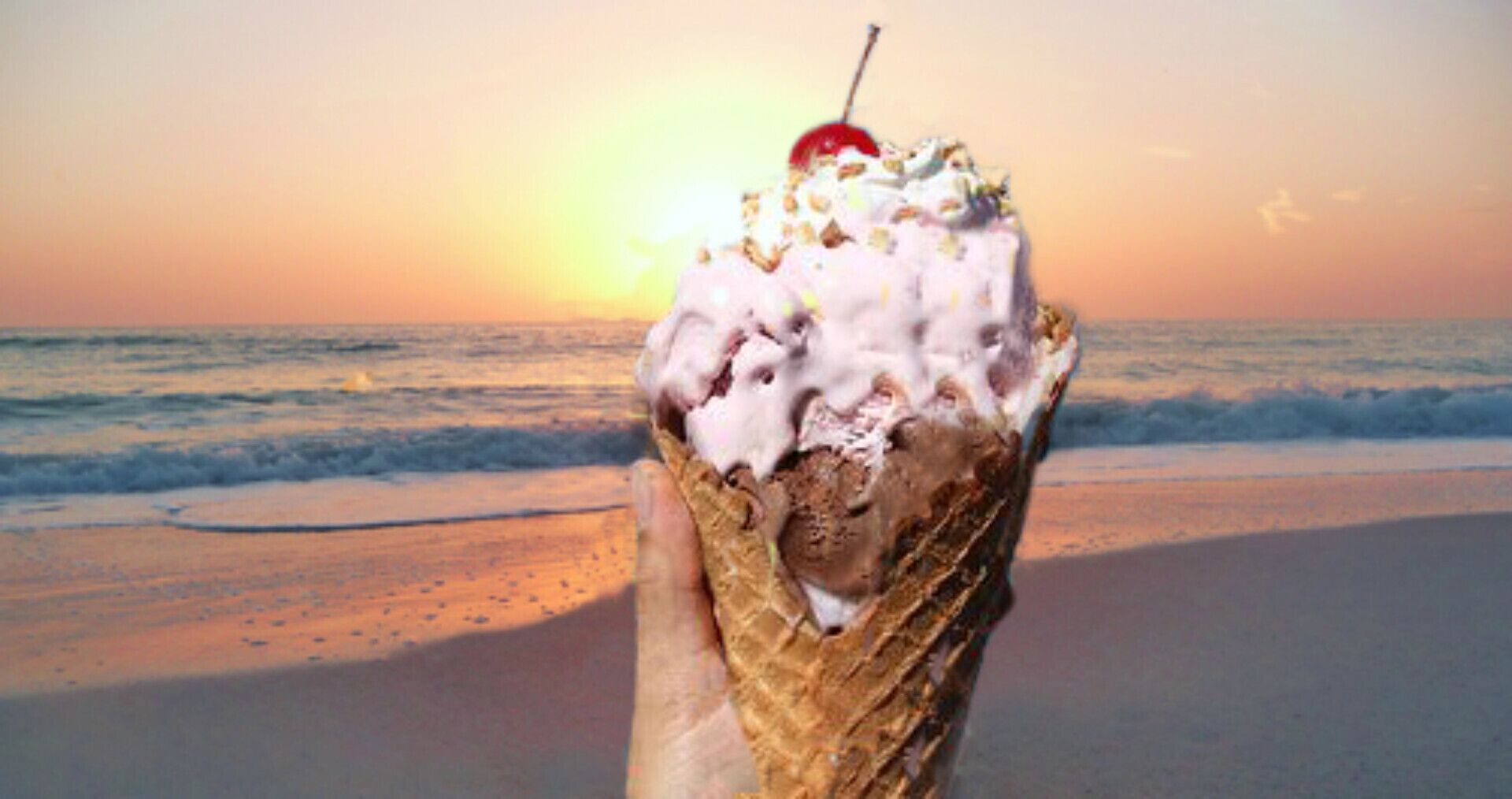 A hand holding an ice cream cone with a cherry on top, set against a beach backdrop with the ocean and a sunset.
